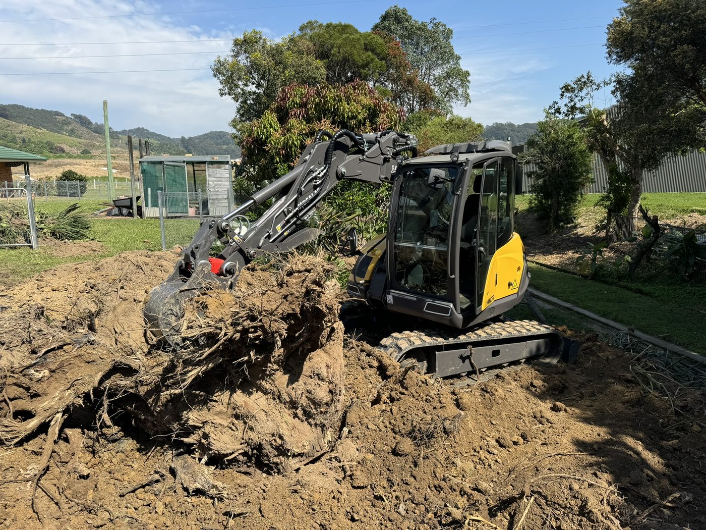 Mecalac excavator lifting a large root ball, stump removal at Roselands Dr Coffs Harbour